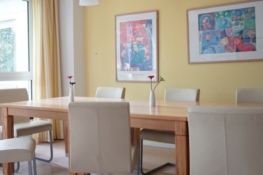 Langer Tisch mit weißen Polsterstühlen vor einer gelben Wand mit bunten Bildern