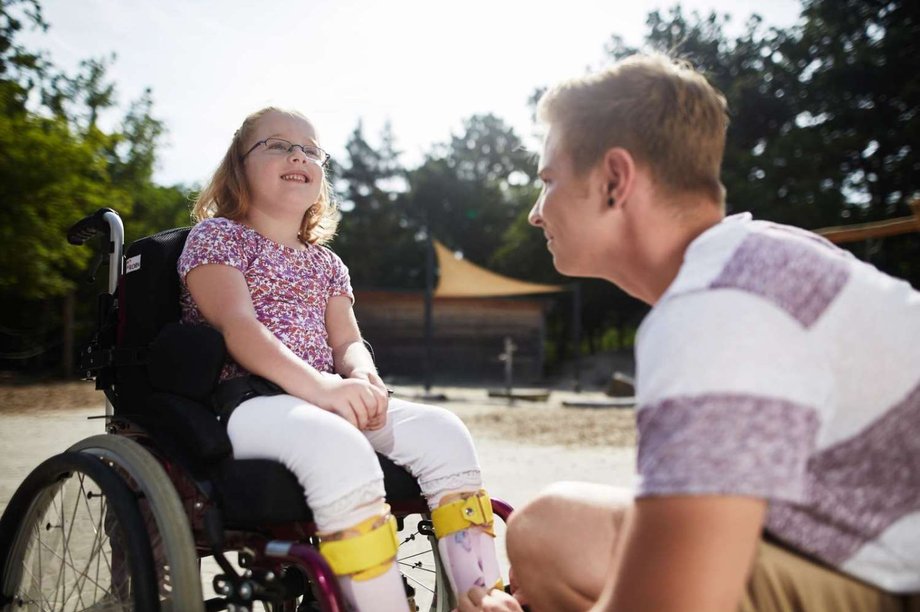 Junger Mann kniet vor einem lachenden Mädchen im Rollstuhl.