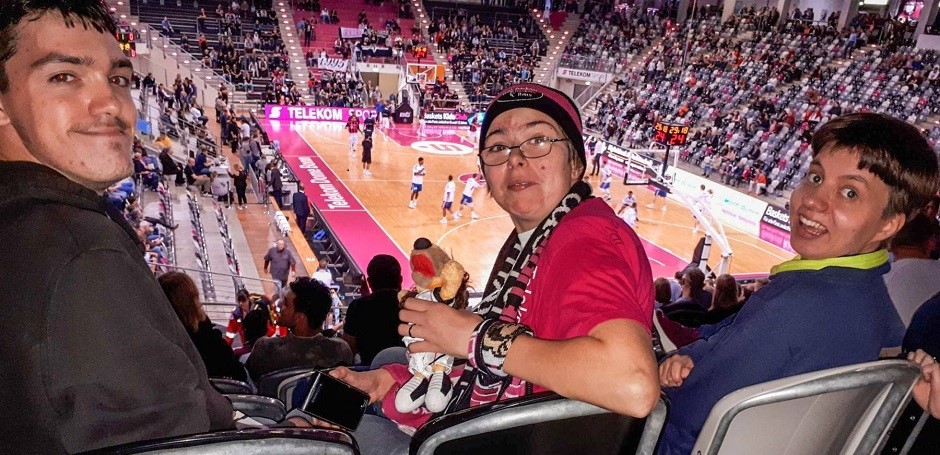 Zwei Frauen und ein Mann sitzen mit Fanbekleidung in einem Sportstadium und schauen sich ein Basketball-Spiel an.